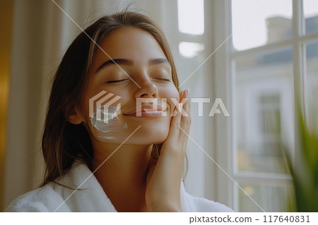 Woman applying facial cream while relaxing indoors near a window in natural light 117640831