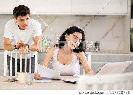 Sad girl works with laptop and paper documents and listens to reproaches of young guy 117830758