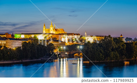 Prague Castle and the Vltava River are illuminated at night, with a small boat rowing across the water in the foreground. The reflections of the lights on the river create a mesmerizing scene. 117989925