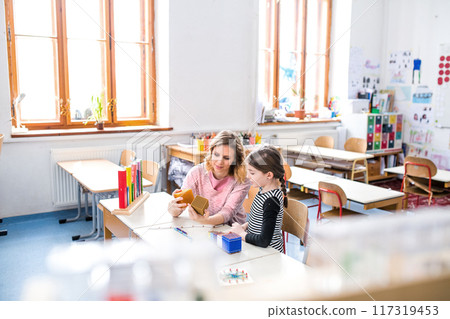 Hardworking teacher learning with young schoolgirl in classroom, explaining math problem, using education tools. 117319453