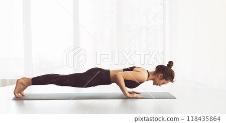 A woman is holding a plank position, engaging her core and muscles in a challenging workout routine. She is focused and determined, showing strength and endurance in her fitness regimen. 118435864