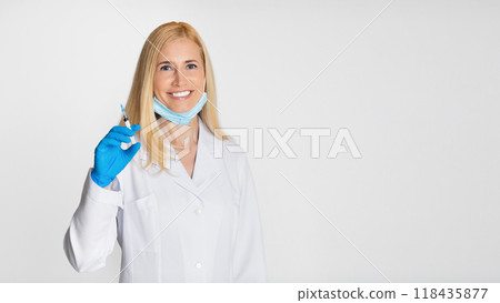 A blonde lady doctor wearing a white lab coat, a light blue face mask, and blue gloves holds a syringe in her right hand and smiles at the camera. The background is a plain white wall. 118435877