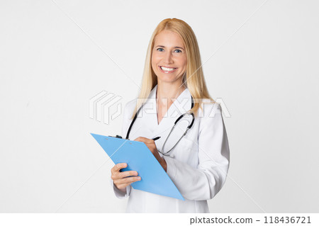 A smiling lady doctor in a white coat with a stethoscope around her neck is writing on a blue clipboard against a white background. She is looking at the camera with a warm and friendly expression 118436721