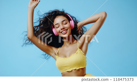 Young woman with wavy dark brown hair dancing joyfully against a light blue background, wearing pink headphones, yellow halter top, and flowy skirt, exuding elegance and joy. Happiness and lifestyle 118475373