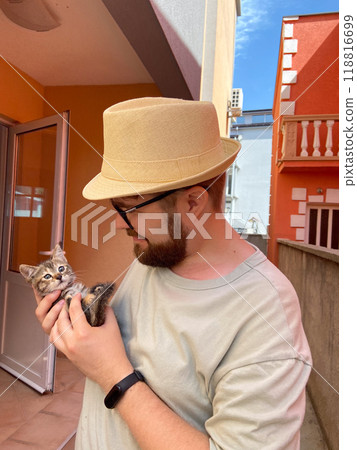 Millennial man in a straw hat and sunglasses holds lovingly at a small kitten in his hands. Tenderness moment captures the bond between human and pet. Themes of care and affection 118816699