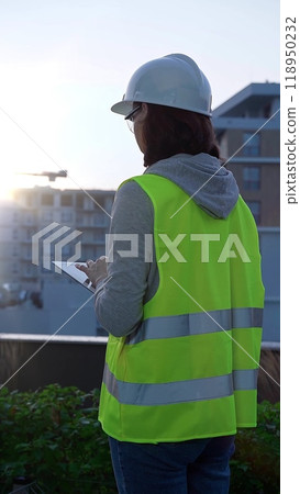 Woman construction engineer with a white hard hat and safety vest is using digital tablet while inspecting a construction site in the early morning at sunrise, back view 118950232