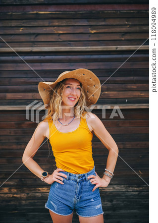 Young woman in a yellow tank top and straw hat poses confidently against wooden backdrop 118094989