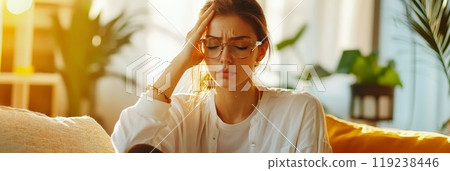 Young woman is sitting on her sofa, experiencing a headache and feeling unwell. She is holding her head with her hand, her eyes closed in discomfort 119238446