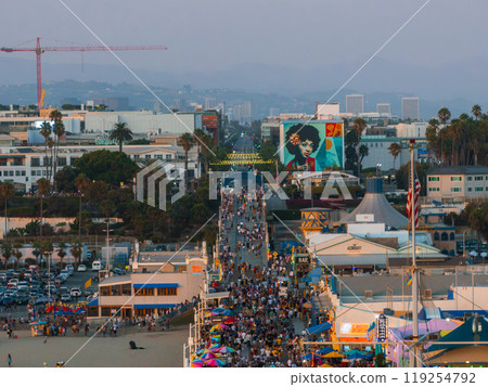 Santa Monica Pier in Los Angeles is crowded with people and vibrant umbrellas. A mural of a person with an afro adorns a building, with palm trees and a crane visible. 119254792