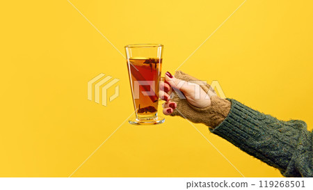 Hand wearing cozy knit mittens holds glass of Zbignew, garnished with cinnamon stick, against warm yellow studio background. 119268501