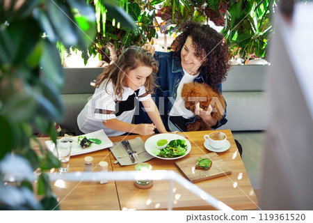 A woman and a girl are seated at a table, enjoying vegetables meal together. Various dishes and utensils are spread out on the table as they engage in conversation and share food 119361520