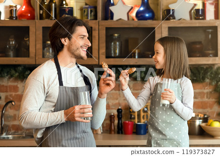 Father and little daughter having a bite in kitchen, eating cookies and drinking milk together, enjoying spending time at home 119373827