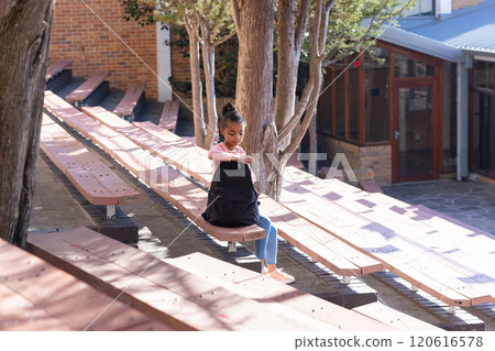 Sitting on bench, african american girl in school uniform looking inside backpack outdoors 120616578