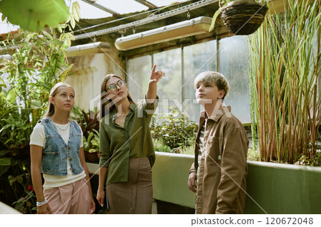 Young Caucasian woman giving tour in botanical garden greenhouse showing plants to teen boy and girl 120672048