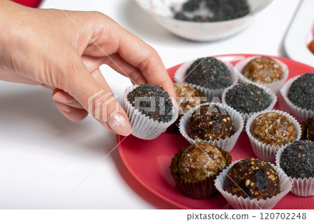 Woman puts dried fruit candies on a plate. Balls of prunes, dates and coconut. With a sprinkle of black sesame powder. 120702248