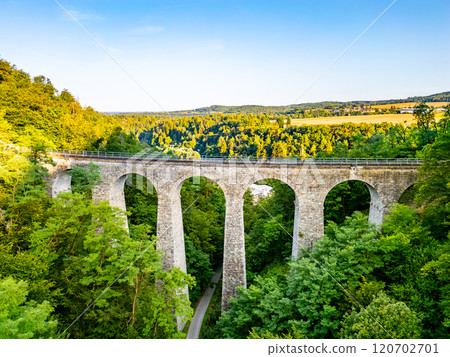 The Zampach stone railway bridge spans lush trees, showcasing impressive stone arches against a clear sky, set in the scenic Czech countryside. 120702701