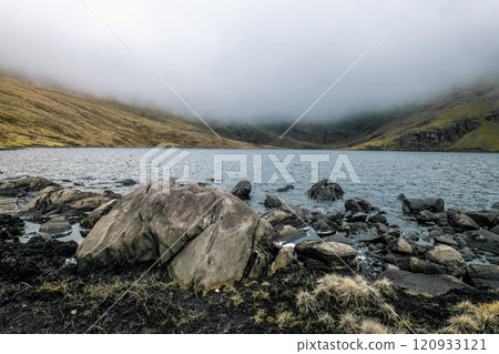 The shores of a lake in a mountainous region, with scattered rocks and fog blanketing the hills. 120933121