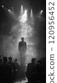 Black and white photo of a man standing in front of a stage in a smoky jazz club 120956452