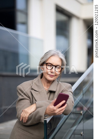 Elderly businesswoman checking her phone while standing outside a modern office building in the 120021285