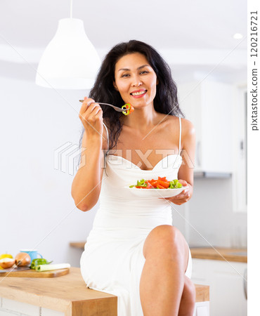 Woman sitting on kitchen table and eating salad 120216721