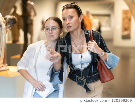 Young woman with teen daughter walk around museum, view sculpture in museum hall 120399269