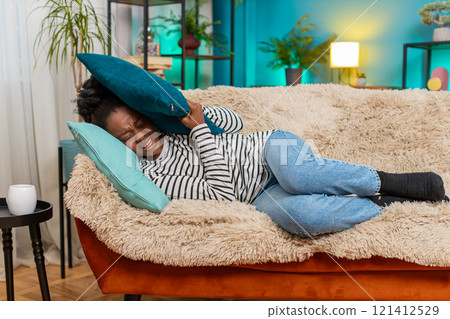 African American woman lying on sofa closing eyes covering face with pillow feeling migraine pain 121412529