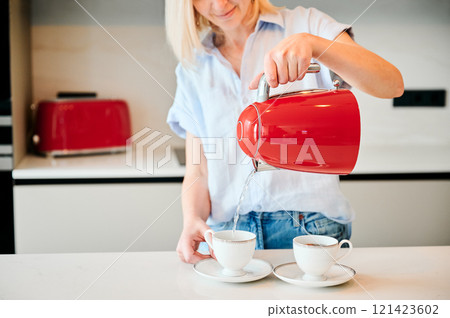 Cropped picture of woman pouring water into cup. Young female making tea for two. Woman holding red kettle over two cups. 121423602