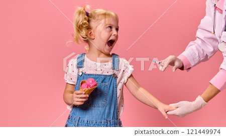 Little girl reacts in shock with open mouth, holding ice cream, hand extended near syringe against pastel pink background. 121449974