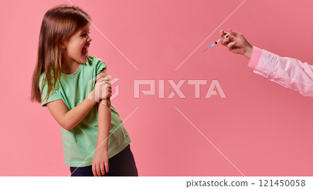 Girl in green shirt reacting dramatically holding hand on arm near syringe held out by doctor against pink studio background. 121450058