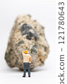 Miniature people construction worker stands next to a massive rock, scratching his head in contemplation 121780643