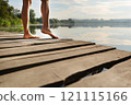 Close-up of bare feet on a wooden dock with a serene lake and trees in the background under a bright sky 121115166