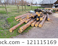 A neat pile of freshly cut logs stacked in a rural yard, surrounded by trees and green grass under a cloudy sky 121115169