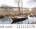 A wooden Viking-style boat resting on a still lake surrounded by reeds and bare trees under a cloudy sky 121115176