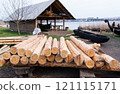 A pile of timber logs near a lakeside workshop, with wooden boats, a barn, and water visible in the background 121115171