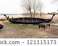 A traditional Viking boat on a grassy shore near the water, with wooden benches nearby and reeds in the background 121115173