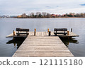 A wooden dock with rustic benches extends over a calm lake, with trees and houses visible in the distance 121115180