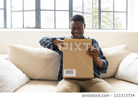 Smiling African american man opening package on couch at home, enjoying holiday surprise 121313734