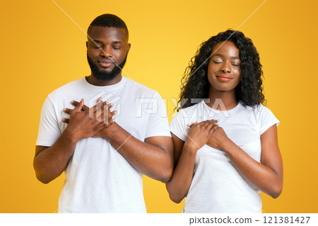 A Black couple stands with their eyes closed, holding their hands over their hearts. 121381427