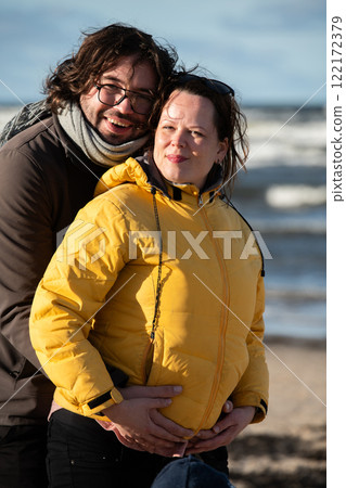 A couple stands embraced on a sandy beach on a chilly autumn day, the serene ocean stretching behind them as they share a quiet moment of love and warmth amidst the cool breeze and peaceful 122172379