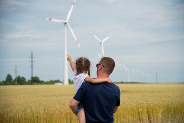 A father holds a young girl as they look towards a wind farm on the horizon