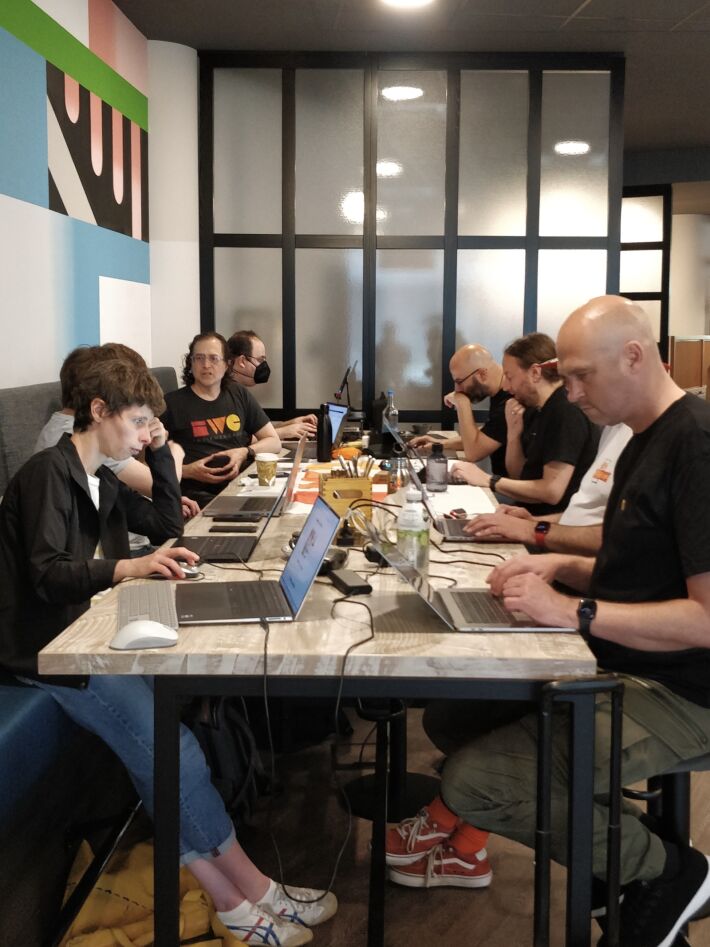 Nine people sitting with laptops at a high table, stools on one side, long couch on the other, typing and having discussions.