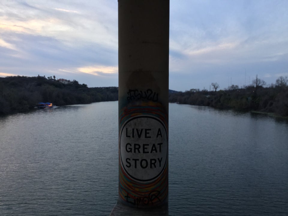 View from a bridge of Lady Bird Lake, with a cement column in front of us dividing the view, with a large white circle painted on it with the all capitals words LIVE A GREAT STORY written inside in a large sans-serif font.