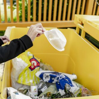 A hand is throwing a plastic food container into a large yellow bin at a household waste collection point.