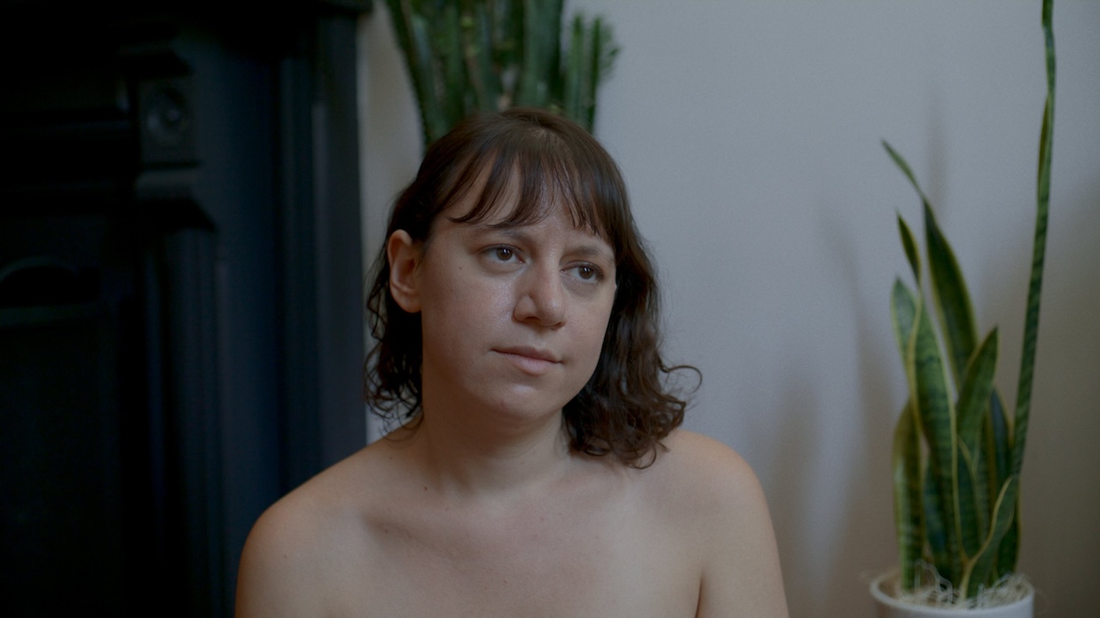 A young white woman with brown hair is topless in front of several potted plants.