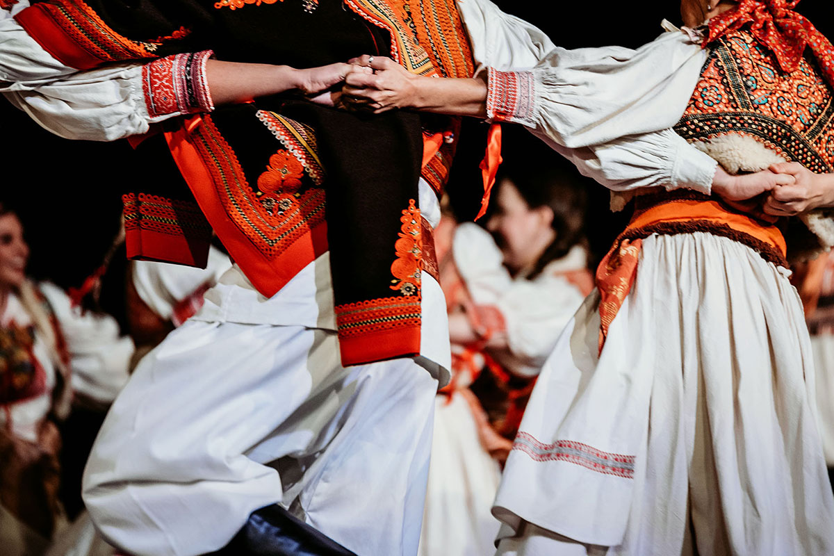 Closeup on the backs of dancers performing in a circle wearing matching white shirts, skirts, and pants and red and black embroidered vests. Each person has their hands around their neighbors' backs, clasping hands with their neighbors on the other side.