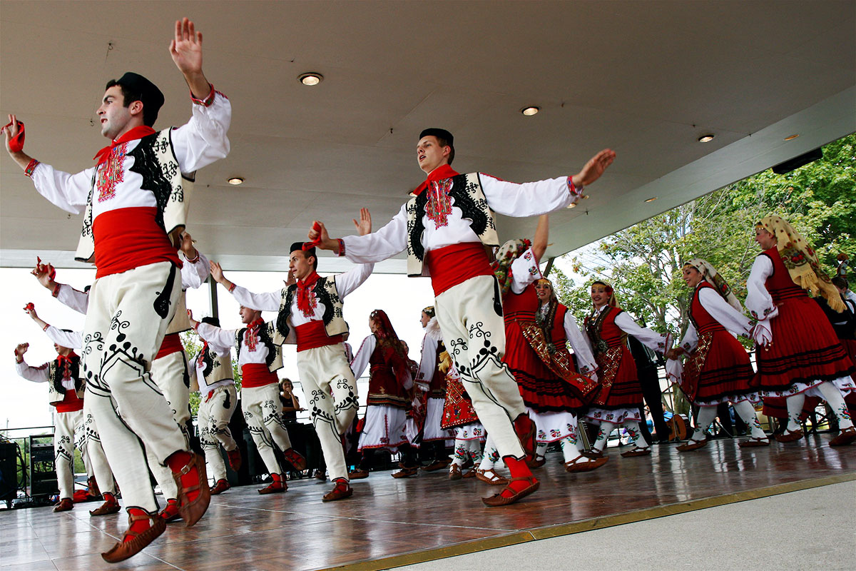 Men and women dance on an outdoor stage, dressing in traditional Croatian costume. Two of the men are mid-jump.