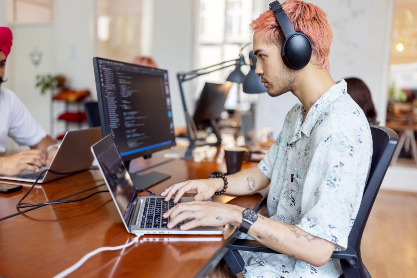 Young man wearing headphones working on computer at startup office. Young IT professional working at coworking office with people working at back.