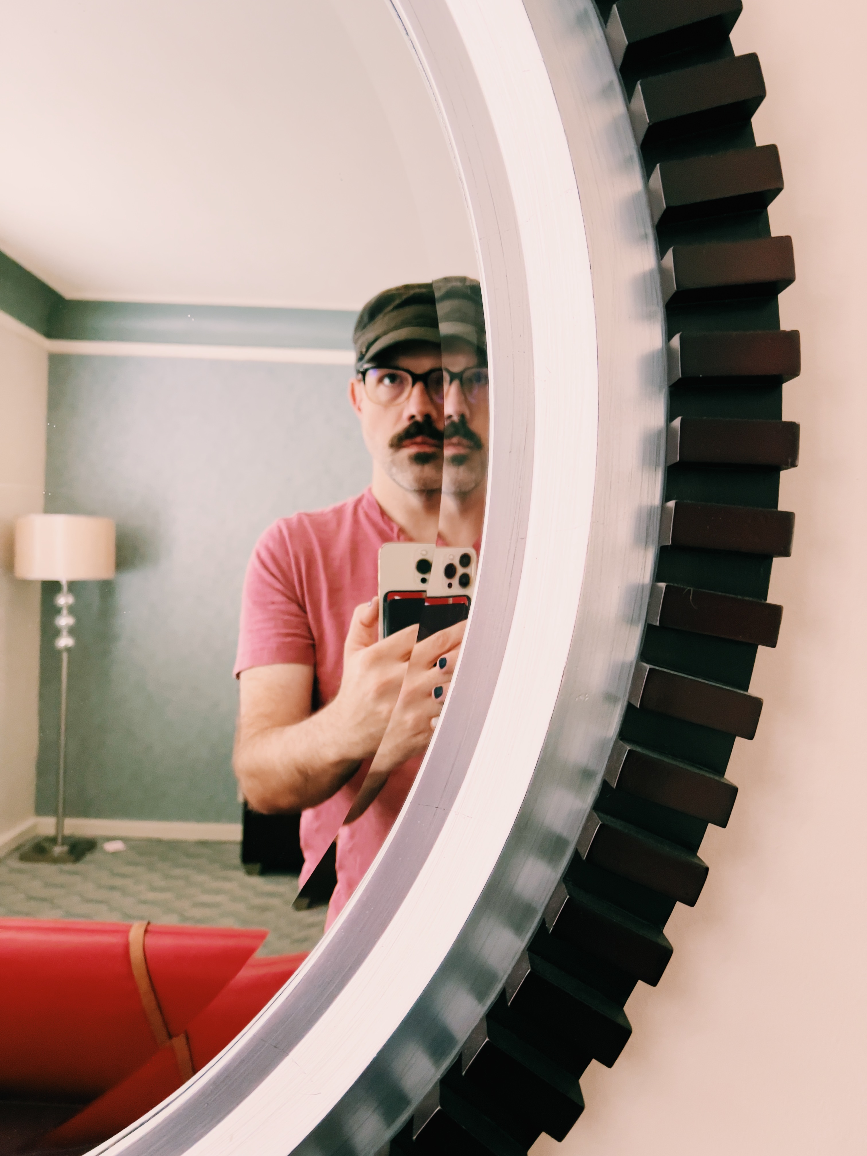 Self-portrait of a masculine-presenting person taking a photo with an iPhone in a mirror. Half of their face is divided by a bezel in the mirror, refracting their face in a disconcerting way. They are wearing a black army cap and a pink henley t-shirt. Their facial hair is a dark, full mustache with a small patch underneath their bottom lip. A small glimpse of dark blue nail polish can be seen on a few of their fingers.