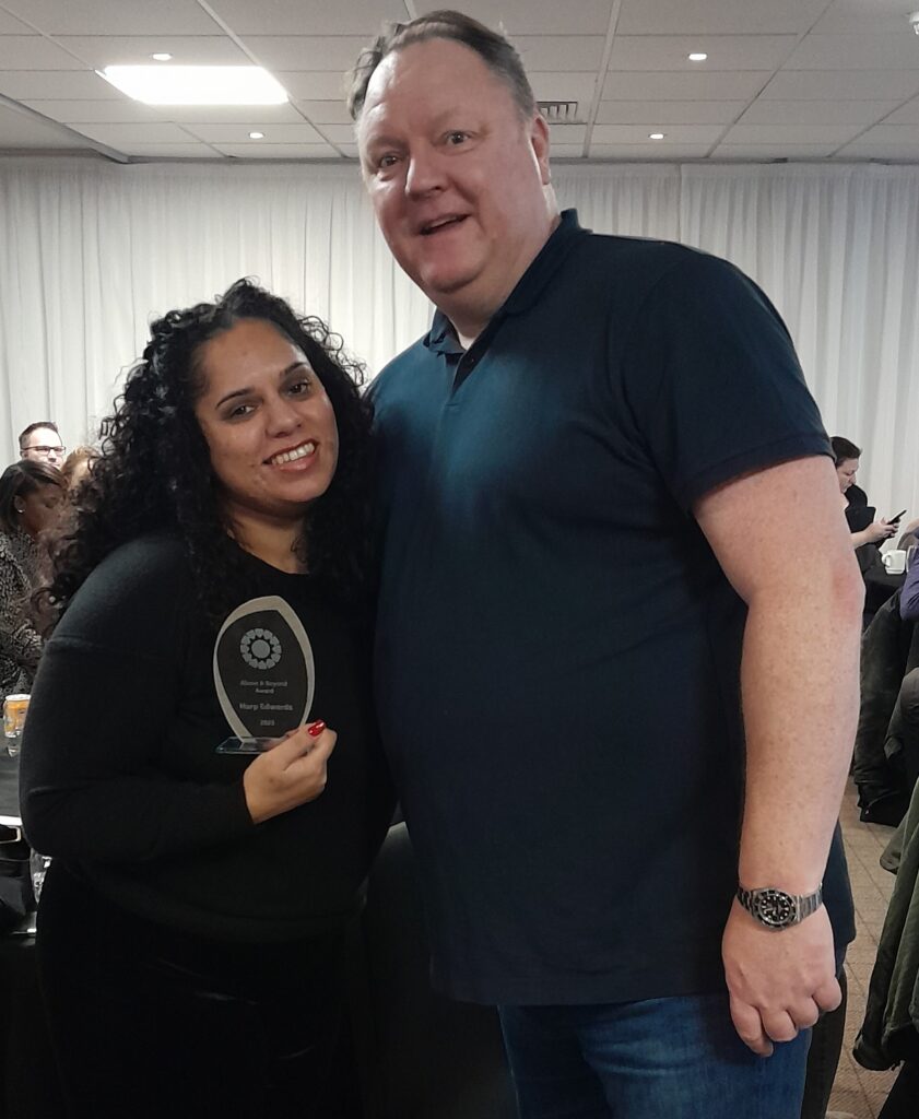 Two people smile toward the camera with one holding a glass award for the Above and Beyond Award at the 2023 Gordon Moody Staff Awards.