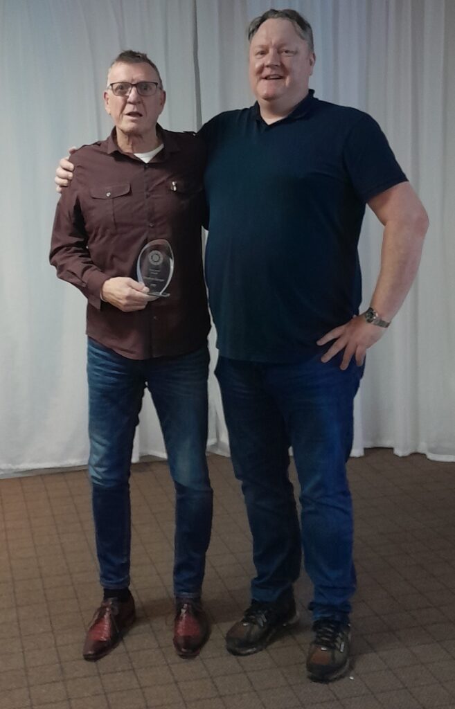 Two people smile toward the camera with one holding a glass award for the Newcomer Award at the 2023 Gordon Moody Staff Awards.